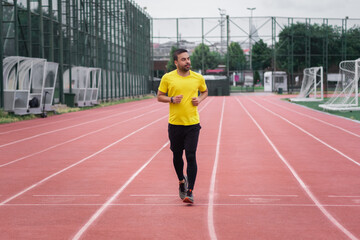 Obraz premium Sportive guy in tracksuit running along red track with white separating lines by soccer field on large sports ground in urban park for health and body development