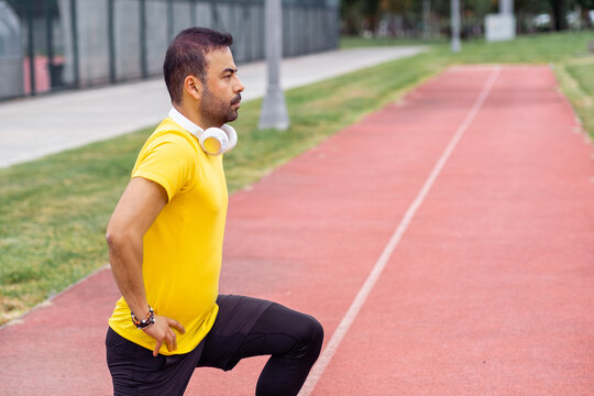 Springtime regular workout: A sportive man in comfortable attire performs squats with hands reaching forward on a running track at an urban park's city stadium. 