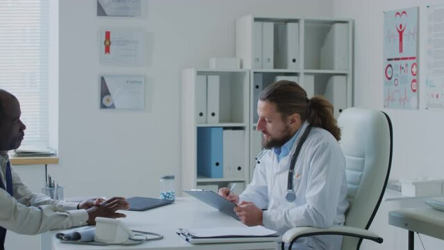 Medium pan shot of Caucasian physician talking to black male patient and writing down complaints about lungpain and cough into medical record during appointment in clinic