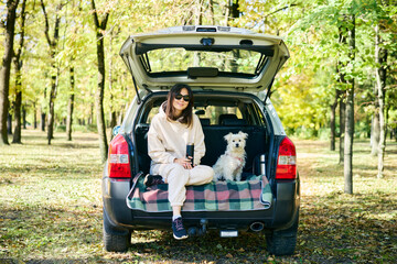 Young woman with a dog relaxing and enjoying nature sitting in car trunk