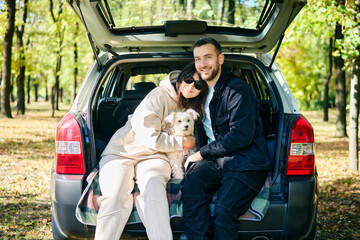 Loving couple embracing hugging with their white dog in the hatchback of a car relaxing in a sunny woodland