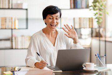 Cheerful middle aged business lady waving hello to laptop indoors