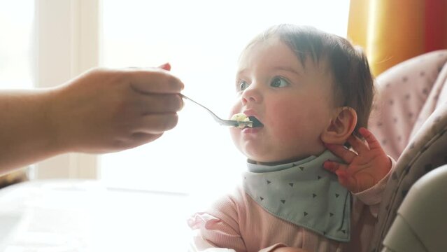 Baby feeding. Parent feed baby porridge. Child sits at table on a chair with a spoon. Happy kid eats porridge with a spoon. The parent feeds the baby with a spoon. Mother nanny takes care of the child