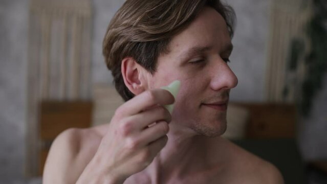 Young man doing facial massage using gua sha scraper. The concept of male cosmetology and care for you and your face