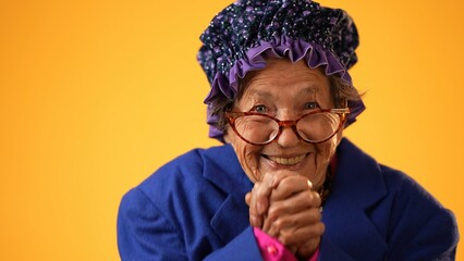 Funny closeup view of smiling happy elderly senior old woman with wrinkled skin smiling with hands together isolated on solid yellow background. - obrazy, fototapety, plakaty