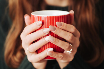 Hot tea in a mug. Woman holding red mug. Cold winter relax background. Green woolen sweater warm clothing. Long hair girl. Cozy atmosphere. Girl hands on coffee mug. Manicure fingernails. - obrazy, fototapety, plakaty