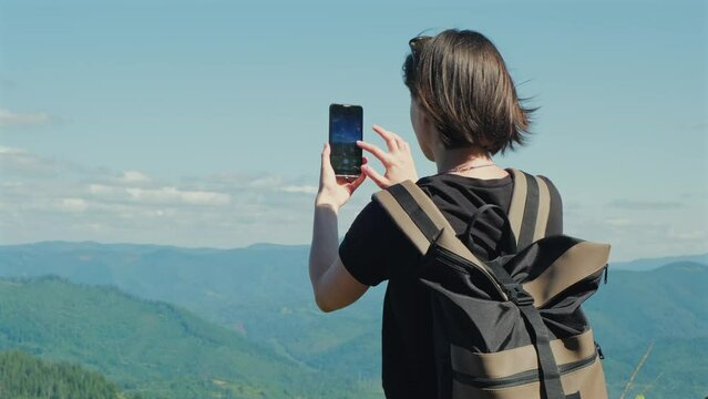 Woman traveler takes photos of mountains landscape on mobile phone. Female hiker travel blogger with backpack creating content for social media outdoors