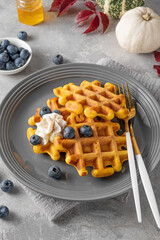 Spiced pumpkin waffles with whipped cream, honey and blueberries on a gray plate on a gray concrete background. Dessert for Thanksgiving Day or Halloween.