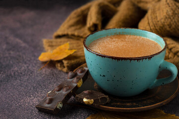 Ceramic blue cup with cocoa, hot chocolate on a saucer, pieces of chocolate with nuts, brown knitted sweater and yellow leaves, autumn composition, horizontal, free space.