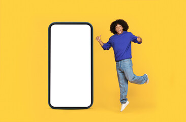 Happy Black Guy Showing Thumbs Up While Jumping Near Big Blank Smartphone