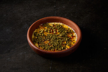 Side view of an elegant crème brûlée with pistachio in a clay bowl, exuding gourmet charm against a stark black backdrop
