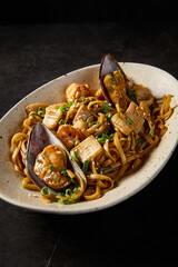 A close-up, vertical side shot emphasizing the rich textures of udon pasta with seafood, curated in Asian tradition, on a ceramic plate over a minimalist black tableau