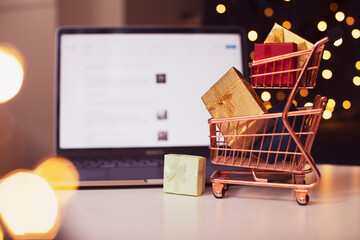 Shopping cart, gift boxes, laptop on the blurred background with bokeh. Christmas shopping online. 