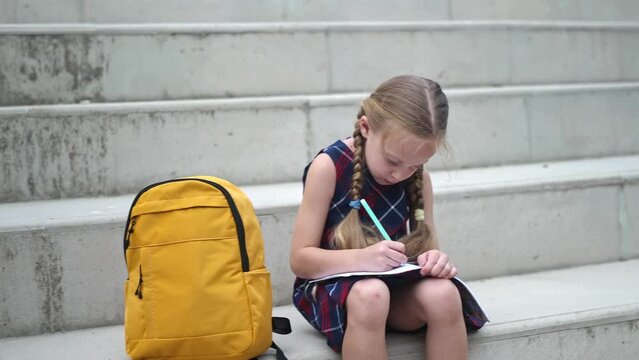 Diligent little girl writes in copybook sitting near backpack on stone steps cute schoolgirl with schoolbag does homework in school yard child does tasks