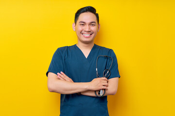 Professional young Asian man doctor or nurse wearing a blue uniform standing confidently while holding stethoscope and crossed hands, smiling friendly at camera isolated on yellow background