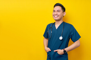 Professional young Asian man doctor or nurse wearing a blue uniform and stethoscope standing confidently while smiling friendly aside, hands in pockets isolated on yellow background - Powered by Adobe