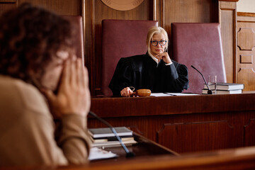Serious blond impartial judge in eyeglasses and mantle looking at young male suspect while sitting...