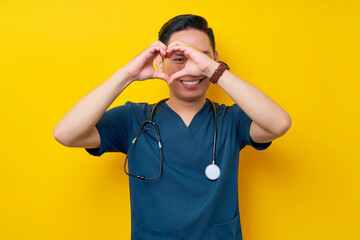 Smiling professional young Asian man doctor or nurse wearing a blue uniform and stethoscope showing heart shape with hands isolated on yellow background. Healthcare medicine concept