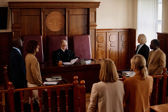 Group of witnesses, attorneys, lawyers, prosecutor and suspect standing in front of experienced blond female judge in mantle passing judgement