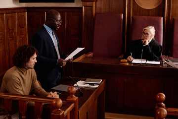 Young confident African American attorney in formalwear reading list of extenuating circumstances while standing in front of judge and prosecutor