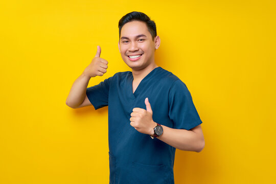 Professional young Asian male doctor or nurse wearing a blue uniform showing thumb up or like, looking at camera with smile isolated on yellow background. Healthcare medicine concept