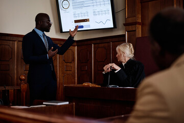 Confident African American lawyer in formalwear pointing at screen with summary of results while explaining data to mature female judge - obrazy, fototapety, plakaty