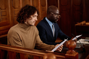 Young male suspect and his attorney looking through juridical documents while sitting in front of camera in courtroom during trial session