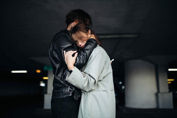 man in a leather jacket and a woman in a coat hugging in an underground parking lot, a couple in...