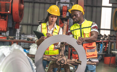 black worker and an Asian female engineer wearing uniform jackets and helmets work inspecting raw materials for steel coils and controlling the production of a roof production line in a factory.