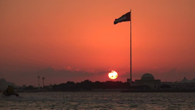 The sun sets over Abu Dhabi behind a flag of the UAE. A man on a jet ski sails towards the sunset.
