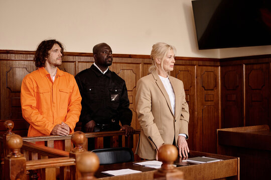 African American Policeman Standing Between Young Male Prisoner In Cuffs And Orange Jumpsuit And Mature Female Attorney During Trial Session