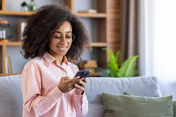 Fototapeta na wymiar Young beautiful woman with curly hair is using the phone, typing messages and browsing online internet pages, sitting at home on the sofa in the living room of the house.
