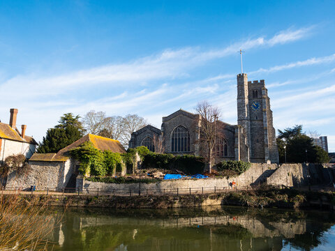 In foreground, the River Medway flows past All Saints Church, a paved riverside walkway and period stone houses at Maidstone, Kent. Bright blue sky, light clouds.