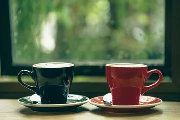  Two cup tea or coffee on wooden table side view window.Red and black hot coffee cup on the table by the window on a rainy day.Side view mugs on glass with reflection in the cafe on vintage style. © arcyto
