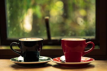 Two cup tea or coffee on wooden table side view window.Red and black hot coffee cup on the table by the window on a rainy day.Side view mugs on glass with reflection in the cafe on vintage style.