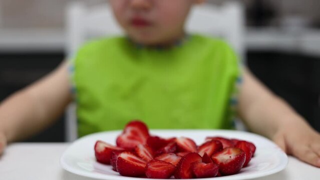 slice of strawberries fruit on plate and baby boy kid is refusing to eat. red strawberry, vitamins healthy nutrition. child with bib in kitchen 4k