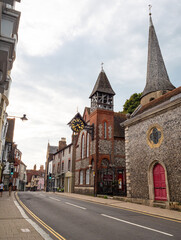 The landmark clock hangs from a tower wall of St Michael in Lewes Church in High Street Lewes, East Sussex, as the street narrows past historic buildings. Clear sky. 