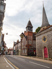 The landmark clock hangs from a tower wall of St Michael in Lewes Church in High Street Lewes, East Sussex, as the street narrows past historic buildings. Clear sky.  © Graham King