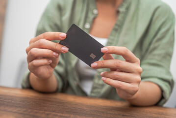 Female examines credit card numbers to enter into online store to make payment through smart system sitting at wooden table in office financial order and purchase