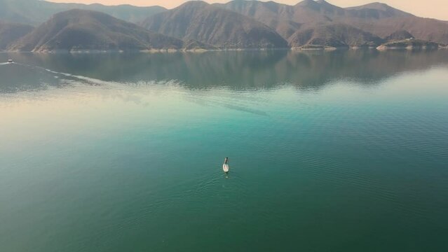 Aerial drone flying over a lake, man doing stand up paddle, with mountains at sunrise. SUP. Beautifull dreamy landscape view. Cosatline with boats. Paceful landscape. Cabra Corral, Salta, Argentina.