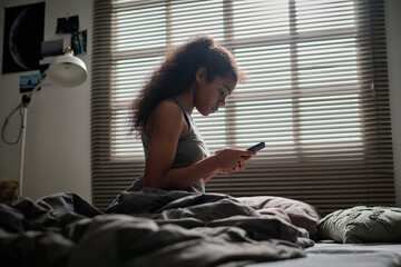 Side view of troubled and sad teenage girl using smartphone while sitting on bed against window...