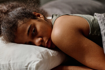 Offended teenage girl in grey vest lying under blanket in bed and keeping head on pillow while...