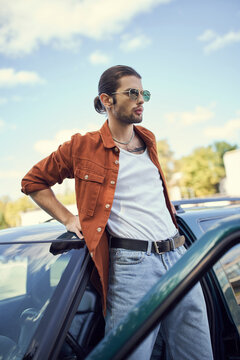 charming young male model in brown shirt and blue jeans standing next to his car and looking away