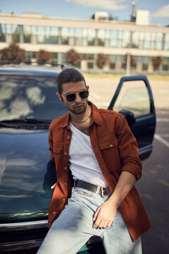 sexy young man in brown shirt and jeans leaning slightly on his car, fashion and style concept