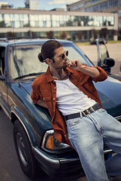 handsome sexy man in vibrant outfit with sunglasses smoking cigarette leaning on his car