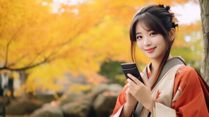 Young Asian woman in traditional kimono stands and smiles while looking at camera with phone on autumn maple leaves at park in Japan