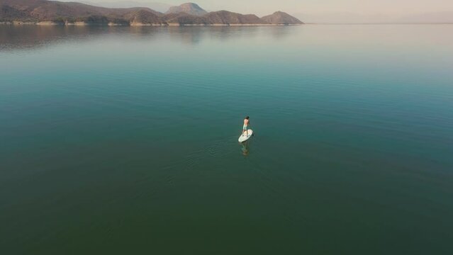 Aerial drone flying over a lake, man doing stand up paddle, with mountains at sunrise. SUP. Beautifull dreamy landscape view. Cosatline with boats. Paceful landscape. Cabra Corral, Salta, Argentina.