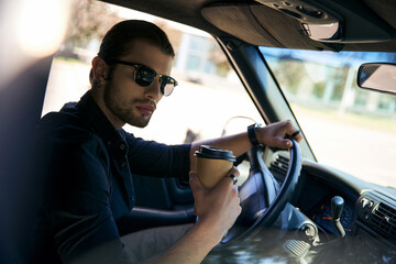 appealing man in black attire holding steering wheel with coffee cup in hand and looking at camera