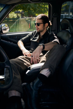 good looking bearded man with tattoos and sunglasses in black outfit sitting in his car, sexy driver