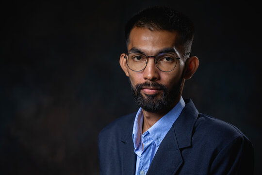 Portrait of young bearded man with glasses and looking at camera on dark background.